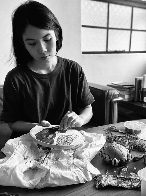 Chong Yi Lin at a table, stitching into a hoop laid over white cloth, with thread, scissors and a pincushion beside her.